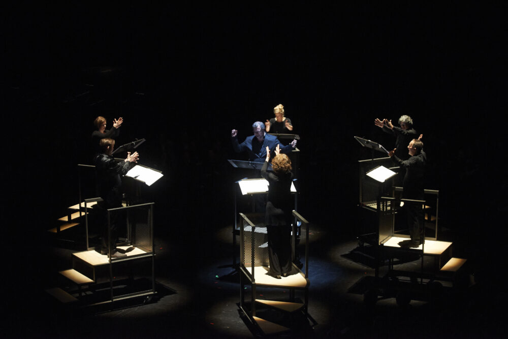 Das Bild zeigt die Neuen Vocalsolisten auf einer Bühne, jede:r auf einem beleuchteten Podest mit Noten. 6 von ihnen bilden einen Kreis, in dessen Mitte der siebte Sänger steht.