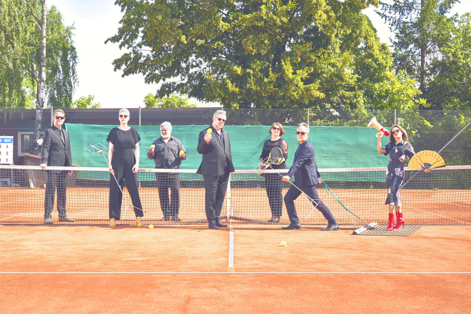 Die sechs Sanger*innen der Neuen Vocalsolisten befinden sich auf einem Tennisplatz. Sie tragen schwarze Kleidung. Ein Mann zieht eine Frau auf dem Schleppnetz. Sie hält ein Megaphon und einen Fächer. Die anderen halten Tennisbälle und Schläger sowie Notenständer in ihren Händen.