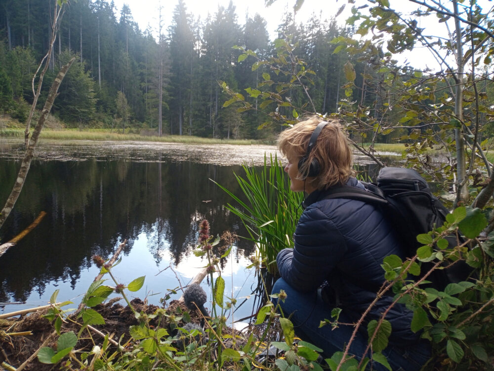 Das Bild zeigt Kirsten Reese. Sie sitzt hochend vor einem See. Im Hintergrund sieht man einen Wald. Sie trägt schwarze Kopfhörer und hat ein Mikrofon vor sich. Auf dem Rücken trägt sie einen Rucksack.