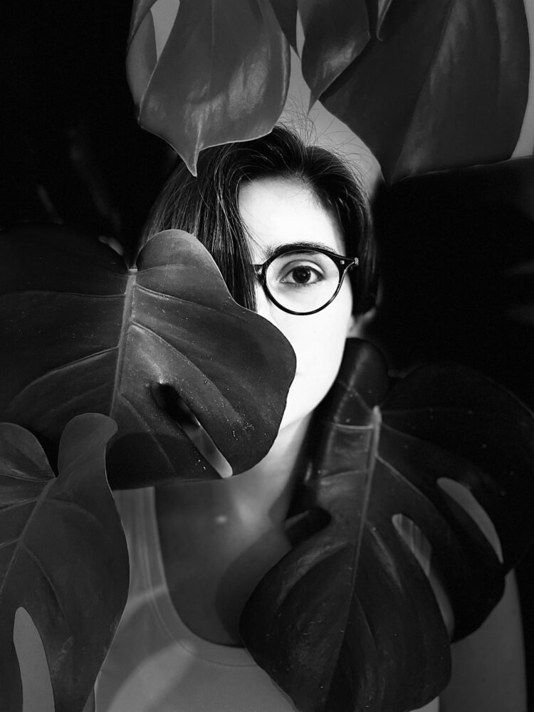 Das schwarz-weißes Porträt zeigt Marta Haladzhun mit dunklen Haaren und runder Brille. Sie ist hinter großen Pflanzenblättern versteckt. Nur ein Auge und Teile des Gesichts sind zwischen den Blättern sichtbar. Die Person trägt dunkle Kleidung und blickt direkt in die Kamera.
