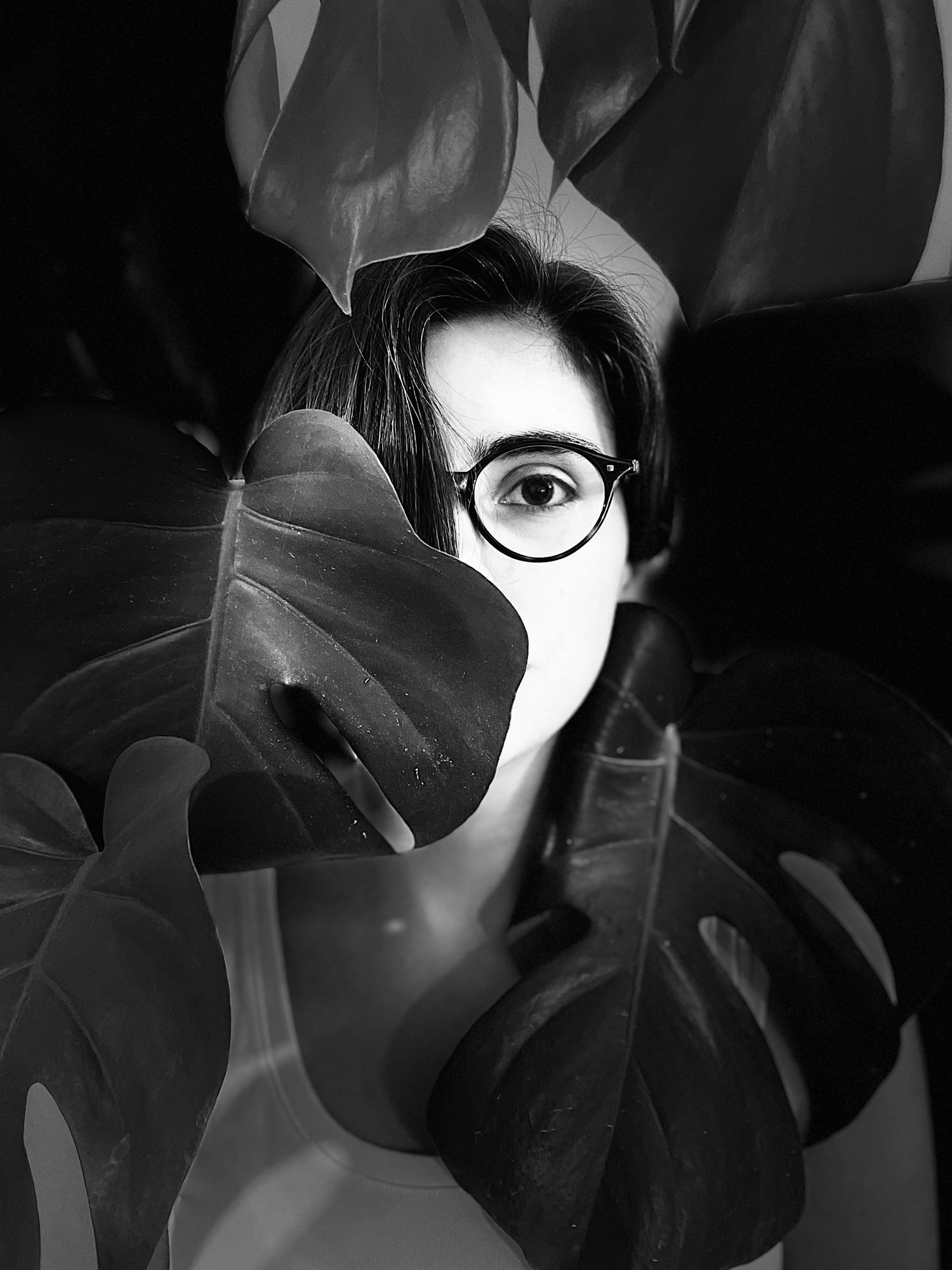 Das schwarz-weißes Porträt zeigt Marta Haladzhun mit dunklen Haaren und runder Brille. Sie ist hinter großen Pflanzenblättern versteckt. Nur ein Auge und Teile des Gesichts sind zwischen den Blättern sichtbar. Die Person trägt dunkle Kleidung und blickt direkt in die Kamera.