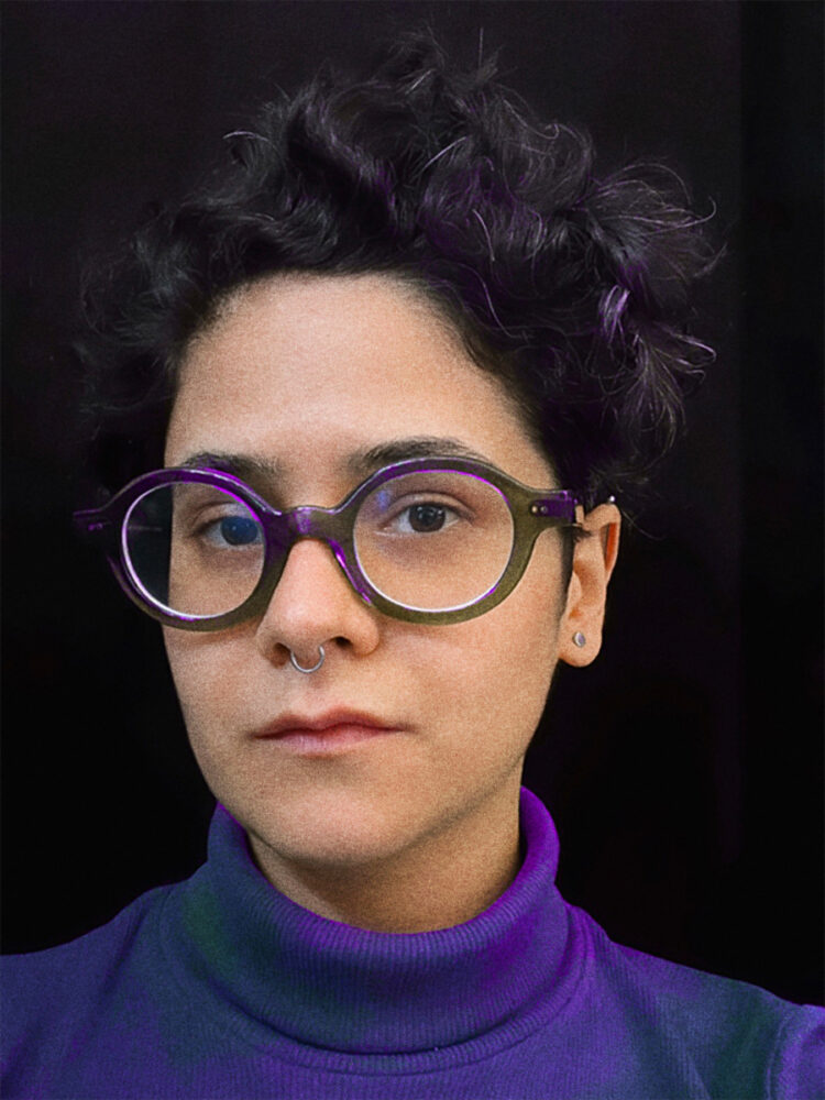 Das Bild zeigt Aida Shirazi mit kurzen, lockigen dunklen Haaren. Sie trägt eine auffällige runde Brille und hat ein Nasenpiercing. Ihr Pullover ist in Blau- und Violetttönen gehalten, auf ihren Haaren und ihrer Brille spiegelt sich violettes Licht.