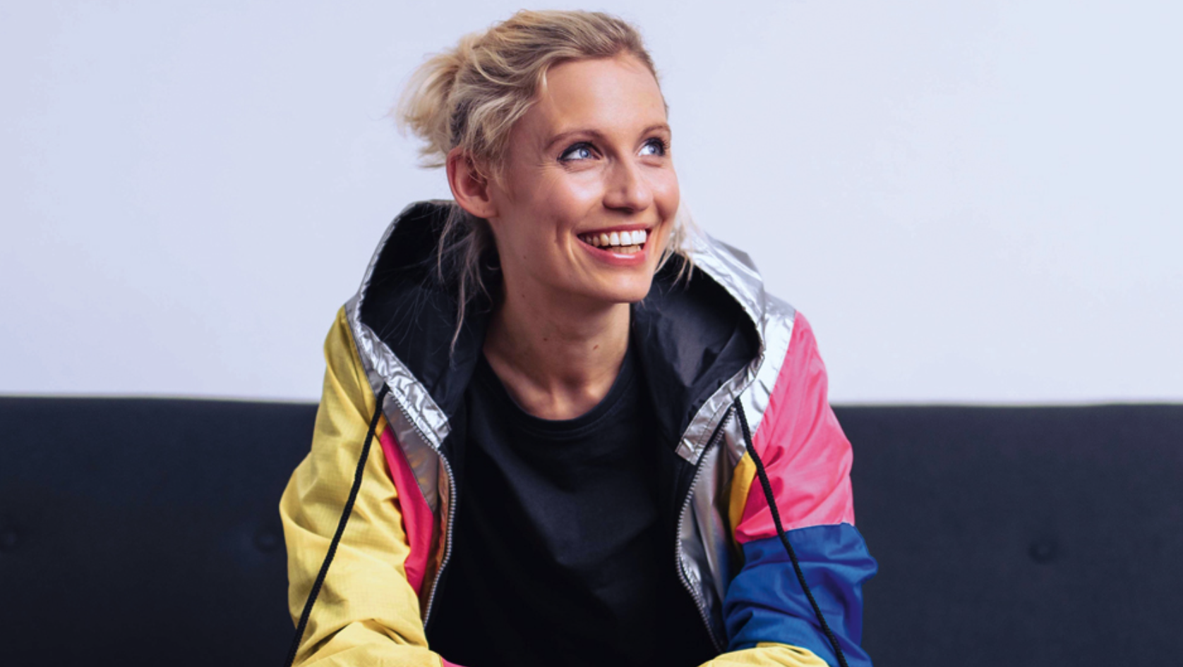 Das Bild zeigt Friederike Scheunchen. Sie trägt einen blonden Zopf und blickt lachend zur Seite. Sie trägt eine bunte Sportjacke in verschiedenen Farben - gelb, pink, blau und silber - über einem schwarzen T-Shirt. Der Hintergrund ist schlicht in Weiß und Grau gehalten.